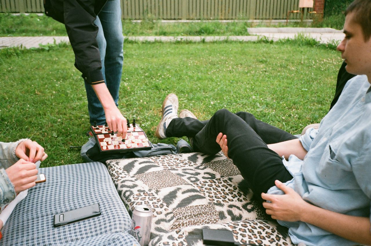 Group of people sitting on blanket in park and playing chess while spending time together on summer day