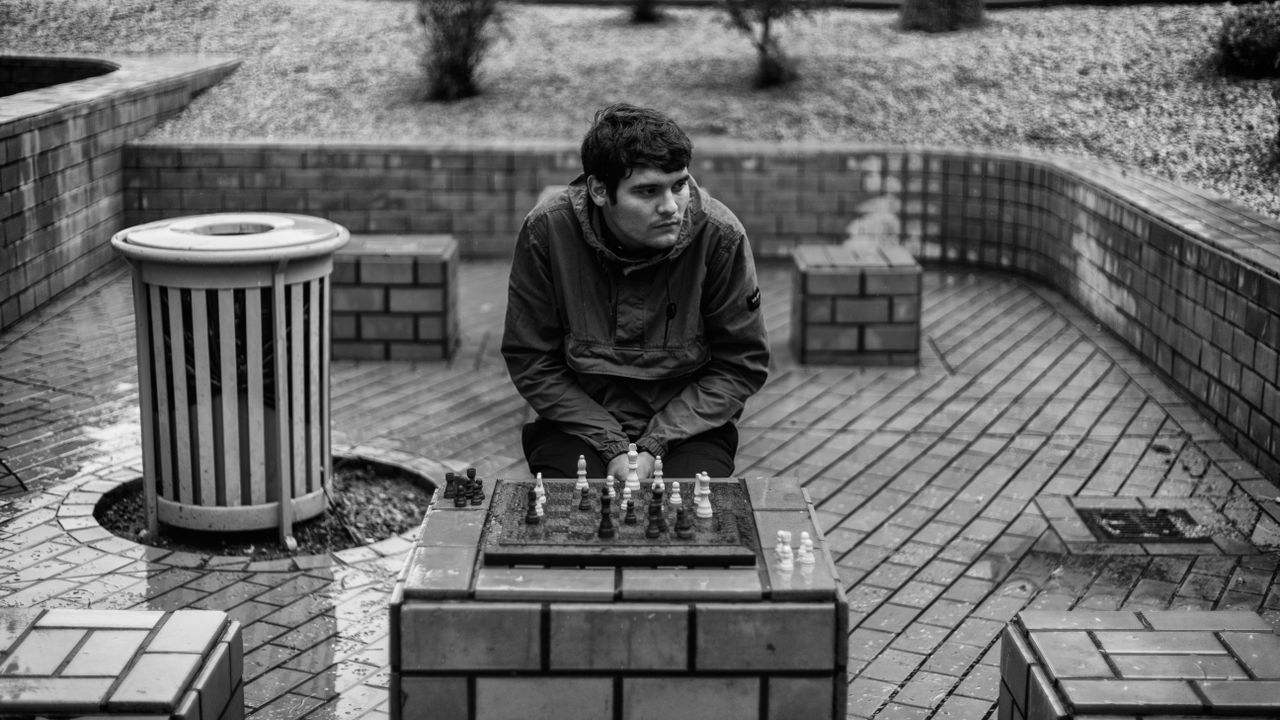 Man engaged in a contemplative chess game in a monochrome urban park.